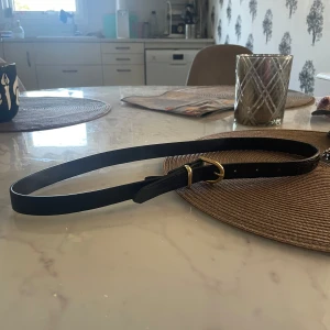 Svart bälte med guldfärgat spänne - Säljer ett stilrent svart bälte med ett snyggt guldfärgat spänne. Bältet har en diskret textur som ger det lite extra karaktär. Perfekt för att piffa upp en outfit, oavsett om det är till vardags eller fest. Passar de flesta storlekar eftersom det är justerbart.