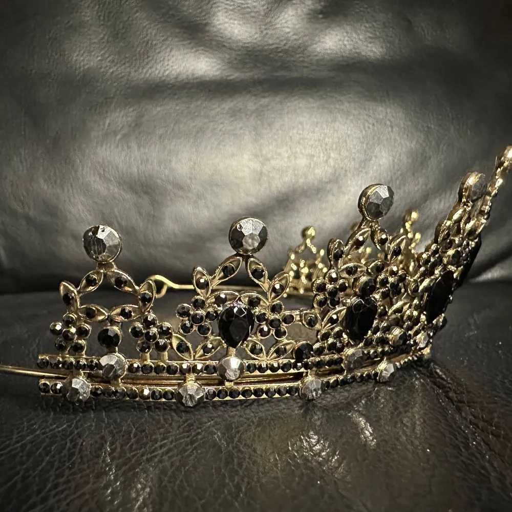 Fantastiskt vacker svart tiara i klassisk modell. Översållad med gnistrande svarta stenar och glittrande svart strass på en mörkt antikguldfärgad botten. Höjd fram 5,5, båglängd 33, bredd 14 cm. Ny och i perfekt skick. Gediget gjord & bekväm att bära. Accessoarer.