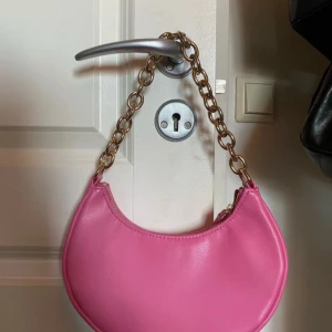 Rosa axelväska med kedjedetalj - Säljer en superfin rosa axelväska med en cool kedja som axelrem. Väskan har en rundad form!  Perfekt för både vardag och fest! 💖pris kan diskuteras vid snabb affär!