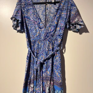 Mönstrad klänning med volangärmar - Säljer en superfin klänning med ett färgglatt paisleymönster i blått, lila och orange. Klänningen har en v-ringad omlott-design och korta volangärmar. Den är perfekt för sommarens fester och har ett knytband i midjan för en snygg passform.