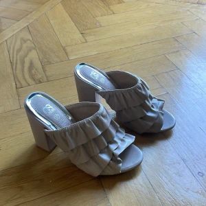 Beige klackskor med volanger - Säljer ett par snygga beige klackskor med volanger. De har en bred klack och öppen tå, vilket gör dem perfekta för sommarens fester. Skorna är i syntetmaterial och har en chic och modern stil.