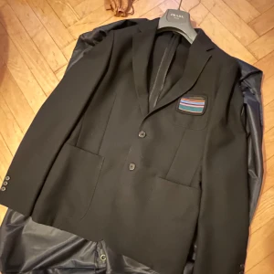 Prada 2018 Kavaj  - Snygg svart kavaj från Prada 2018 med en unik broderad detalj på bröstet. Kavajen har två knappar framtill, 3 yttre fickor och innerficka. kavajen är i mycket gott skick och kommer med galje och dustbag. 