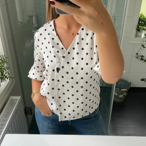 Vit blus med svarta prickar - Säljer en superfin vit blus med svarta prickar. Den har en omlott-design och korta ärmar, vilket gör den perfekt för både vardag och fest. Blusen är i ett lätt och luftigt material, vilket gör den skön att bära under varmare dagar. Passar perfekt till jeans eller kjol!
