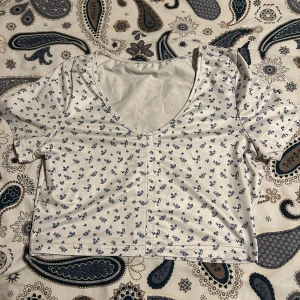 Blommig t-shirt - En vit t-shirt med blåa blommor på. Urringad, säljer pga att den inte kommer till användning. En röd färg klick på baksidan men inget som märks så mycket.