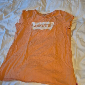 Orange t-shirt från Levi's - Säljer en snygg orange t-shirt från Levi's i storlek M. Den har en klassisk rund hals och korta ärmar. T-shirten är i bomull och har det ikoniska Levi's-loggan tryckt i vitt på framsidan. Perfekt för en avslappnad stil!