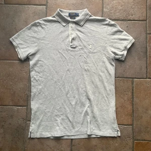 Grå pikétröja från Ralph Lauren - Säljer en stilren grå pikétröja från Ralph Lauren i storlek M. Tröjan har en klassisk krage och knappar framtill. 