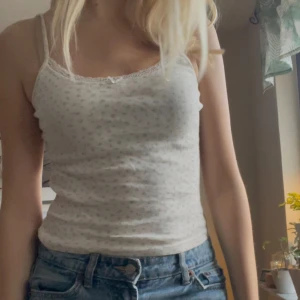 Vit blommig linnetopp - Säljer en söt vit linnetopp med små blommor och spetsdetaljer vid halsringningen. Perfekt för sommardagar eller som ett lager under en kofta. Toppen har smala axelband och en tight passform.