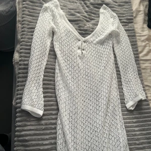Vit stickad klänning - Säljer en superfin vit stickad klänning med v-ringning och knappar framtill. Den har långa ärmar och ett snyggt, luftigt mönster som passar perfekt för både vår och sommar. Klänningen är i nyskick och passar även perfekt till stranden.