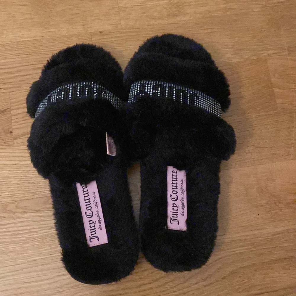 Säljer ett par supermysiga svarta tofflor från Juicy Couture. De är täckta i fluffigt material och har en glittrig rem med texten 'JUICY' i strass. Perfekta för att hålla fötterna varma och stiliga hemma. Innersulan är också klädd i mjukt material.. Skor.