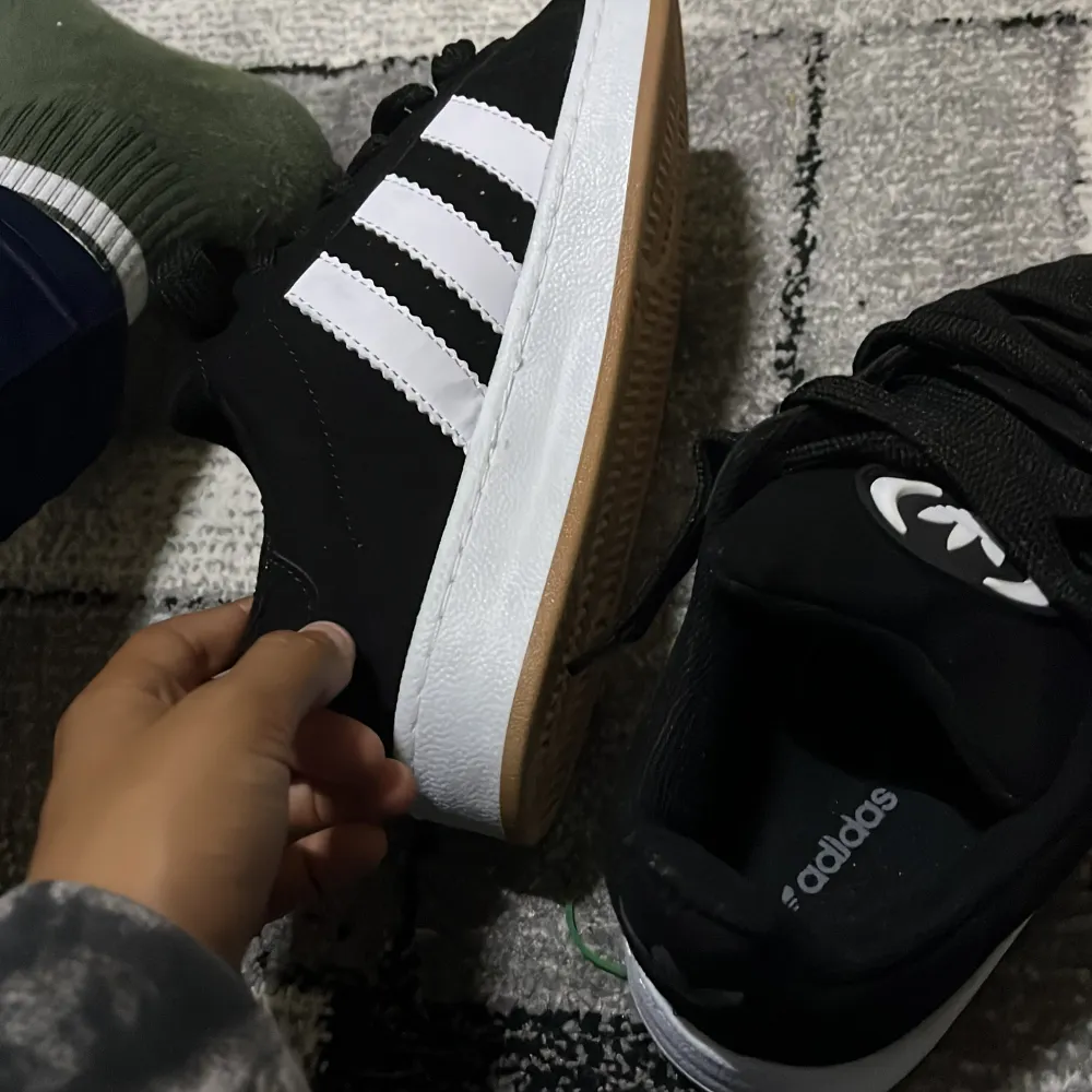 Säljer ett par svarta Adidas Campus sneakers i nyskick. Skorna har den klassiska tre-randiga designen i vitt och är gjorda i skinn. De kommer med extra vita skosnören. Perfekta för både vardag och fest!. Skor.