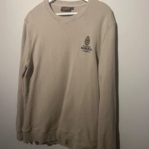 Beige tröja från Morris - Säljer en snygg beige tröja från Morris i storlek L. Den har en klassisk design med rund hals. Perfekt för en stilren och avslappnad look. Morris-loggan är broderad på bröstet för en extra touch av stil.