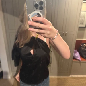 Svart topp med guldkedja - Säljer en snygg svart topp med en unik guldkedja som axelband. Toppen har en stilren design med en liten öppning framtill. Perfekt för en utekväll eller fest! 💫