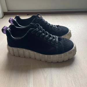 Säljer ett par svarta sneakers från eytys med en riktigt cool och chunky vit sula. De har snörning och en lila flik baktill för extra stilpoäng. Perfekta för både vardag och fest!