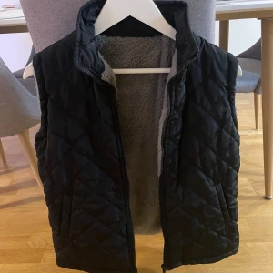 Svart quiltad väst med dragkedja - Snygg svart quiltad väst med dragkedja och mjukt foder. Perfekt för kyligare dagar och ger en stilren look. Västen har en hög krage och praktiska fickor.