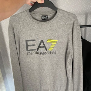 Grå långärmad tröja från Emporio Armani - Säljer en stilren grå långärmad tröja från Emporio Armani med EA7-logga i svart och grönt på bröstet. Perfekt för en avslappnad och trendig look. 