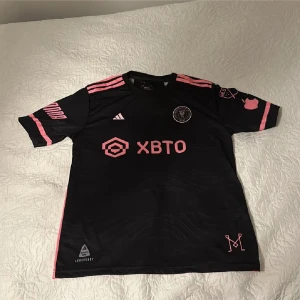 Svart och rosa fotbollströja - Snygg svart fotbollströja med rosa detaljer från Adidas. Tröjan har korta ärmar och klubbmärke på bröstet. Den har även sponsorlogga och spelarnamn samt nummer 10 på ryggen. Perfekt för fotbollsfans!