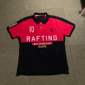 Röd och svart pikétröja från Ralph Lauren - Säljer en snygg pikétröja från Ralph Lauren i rött och svart med texten 'RAFTING' och 'WHITEWATER GUIDE' på framsidan. Tröjan har korta ärmar och en klassisk krage med knappar. Perfekt för en sportig look! Tröjan passar mer som large 