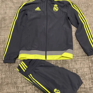 Real madrid Dress - Säljer en grå träningsdress från Adidas med gula detaljer och Real Madrid-logga. Jackan har dragkedja och långa ärmar med gula ränder längs sidorna. Perfekt för fotbollsträning eller som sportig vardagsdress.