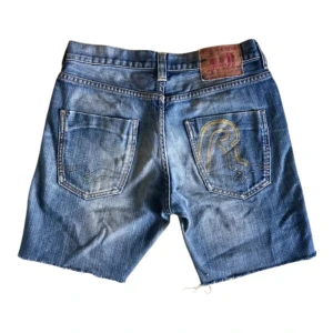 Vintage Blå Replay jeansshorts med råa kanter - Säljer ett par coola. slitna blå jeansshorts med råa kanter. Vintage från 2000. De har en klassisk femficksdesign och en knappgylf. Storlek 36 (barn).