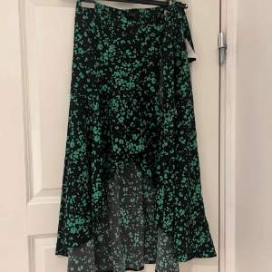 Snygg svart kjol med grönt mönster. Kjolen har en omlott-design och knytning i midjan för en justerbar passform. Perfekt för att ge en färgklick till din outfit! Helt oanvänd med prislapp. Volanger. 