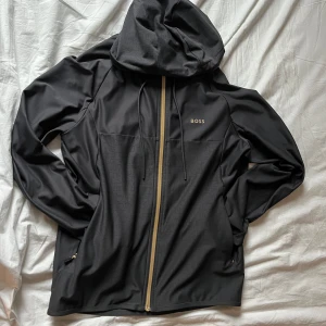 Tunnare mörk grå hoodie med mönster från Boss - Snygg svart hoodie från Boss med ett subtilt geometriskt mönster på framsidan och huvan. Jackan har en dragkedja i kontrasterande färg och är tillverkad i ett snabbtorkande material. Perfekt för en stilren look. Nypris 1799:- SEK 