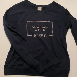 Mörkblå tröja från Abercrombie & Fitch - Säljer en stilren mörkblå tröja från Abercrombie & Fitch med tryck på framsidan. Tröjan har långa ärmar och en rund halsringning. Perfekt för en avslappnad stil. Strl M men passar mig med S perfekt 