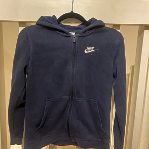 Mörkblå hoodie från Nike - Säljer en mörkblå hoodie från Nike med dragkedja och huva. Tröjan har långa ärmar och två fickor framtill. Perfekt för en avslappnad stil. 🌀