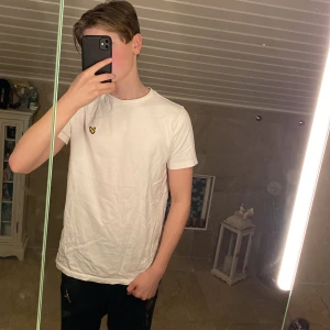 Vit t-shirt från Lyle & Scott - Säljer en stilren vit t-shirt från Lyle & Scott med ett broderat gult emblem på bröstet. T-shirten är kortärmad och tillverkad i bomull, perfekt för en avslappnad look. Storlek: 15 år. 10/10 skick, bara testat. Den kommer även ifrån Jeansbolaget🤙