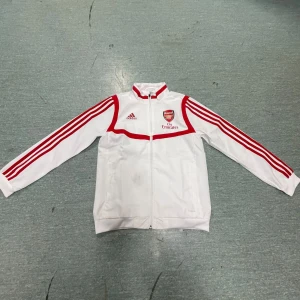 Vit Adidas jacka med Arsenal-logga - Säljer en vit Adidas jacka/hoodie med röda detaljer och Arsenal-logga. Jackan har dragkedja och långa ärmar med klassiska Adidas-ränder. Perfekt för fotbollsfans! 🏆lite smutsig annars inget fel