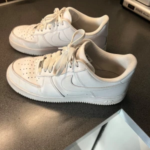 Vita Nike Air Force sneakers - Snygga vita Nike Air Force sneakers med klassisk design och snörning. Perfekta för en stilren look. Skorna har en ikonisk Nike-logga på sidan och AIR-text på hälen.