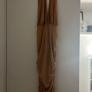 Beige långklänning - Elegant beige långklänning med djup v-ringning och draperad design. Perfekt för en stilfull kväll. Klänningen har en smickrande passform och är tillverkad i ett mjukt material som ger ett vackert fall.