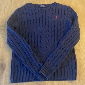 Blå stickad tröja från Polo Ralph Lauren - Säljer en snygg blå stickad tröja från Polo Ralph Lauren med kabelstickat mönster och röd logga på bröstet. Tröjan har långa ärmar och ribbade muddar vid ärmslut och nederkant. Perfekt för kyligare dagar!