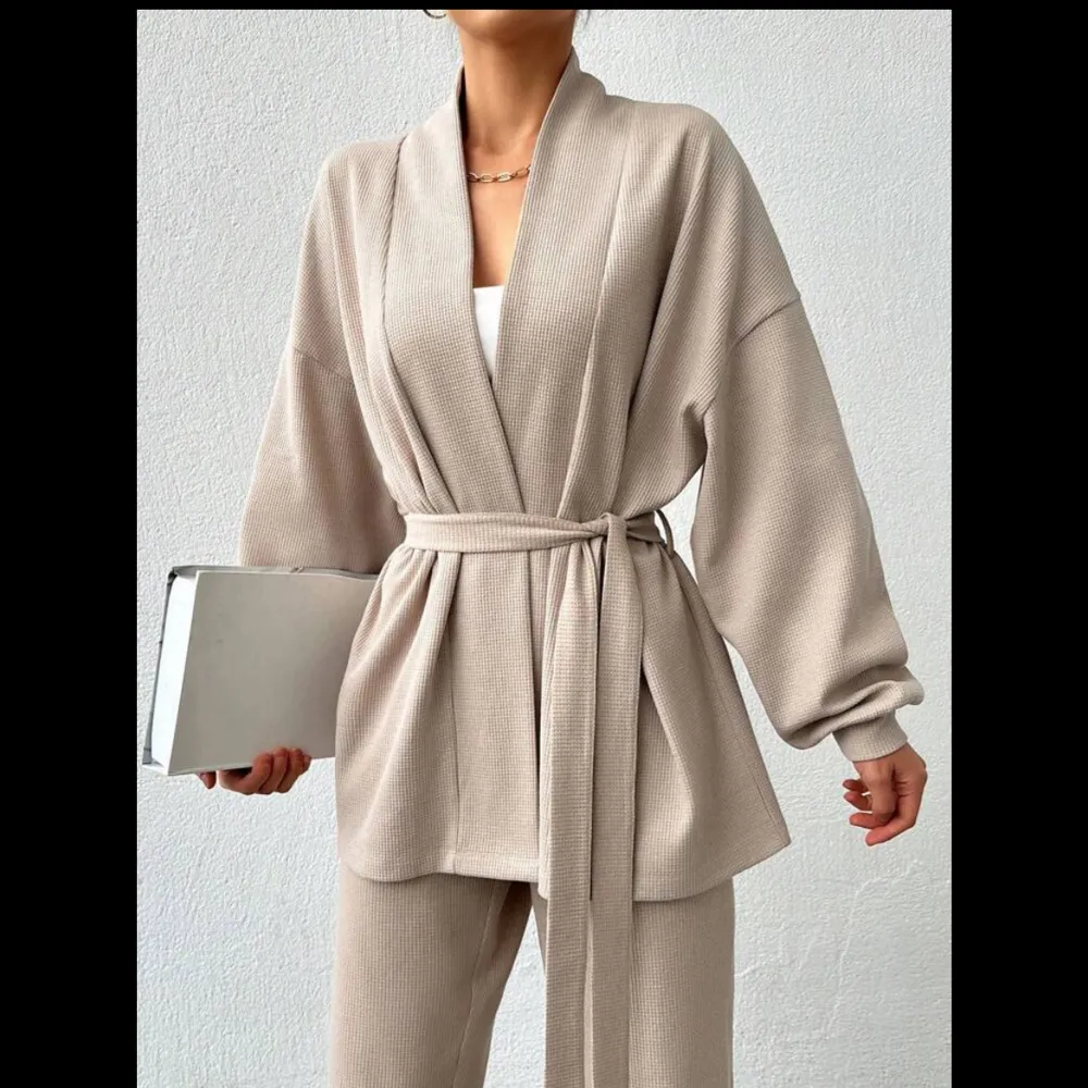 Stilrent beige set med en kofta och matchande byxor. Koftan har en knytning i midjan och vida ärmar, vilket ger en elegant look.. Puvut & Bleiserit.