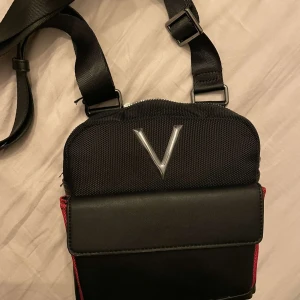 Svart axelväska från Valentino - Snygg svart axelväska från Valentino med justerbar rem och stilren design. Väskan har en framficka med lock och en dragkedja på baksidan för extra säkerhet. Perfekt för att bära dina nödvändigheter med stil.