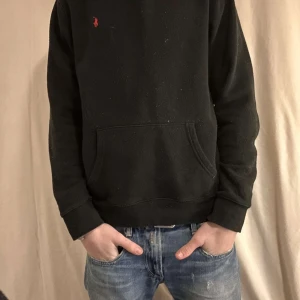 Svart hoodie från Ralph Lauren - Säljer en stilren svart hoodie från Ralph Lauren med en liten röd logga på bröstet. Tröjan har en klassisk känguruficka och långa ärmar. Perfekt för en avslappnad look. Liten vit fläck på magen 