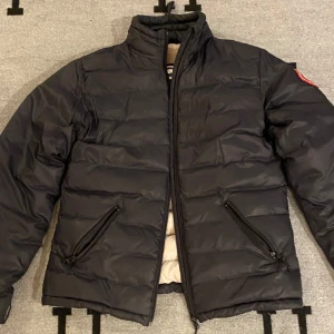 Svart dunjacka från Canada Goose - Säljer en svart dunjacka från Canada Goose med broderad logga på ärmen. Jackan har dragkedja framtill och två sidofickor med dragkedjor. Insidan är ljus och quiltad för extra värme. Perfekt för kyliga dagar!