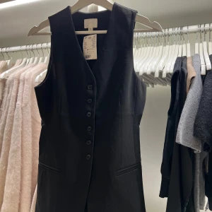 Kostymväst från H&M - Svart väst med knappar framtill. Perfekt för en stilren look. Västen har en design med en v-ringad hals och är från H&M