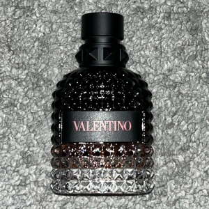 Valentino Born in Roma Uomo Coral Fantasy - Elegant parfymflaska från Valentino med en lyxig design. Flaskan är mörk med en glittrande yta och har en svart kork med nitar. Perfekt för den som söker en sofistikerad doftupplevelse.