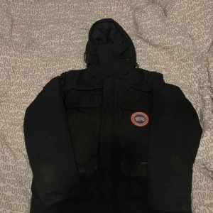 Svart dunjacka från Canada Goose - Säljer en svart dunjacka från Canada Goose med huva och broderad logotyp på bröstet. Jackan har långa ärmar och är perfekt för kalla vinterdagar. Några små hål i den men inget super allvarligt. Den är tillverkad i polyester och fylld med gås fjädrar för extra värme. Storlek Xl