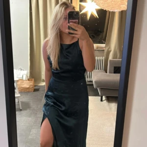 Mörkgrön satin klänning -  mörkgrön klänning i satin med en slits och öppen rygg. Perfekt för speciella tillfällen, tex bal . Den har en smickrande passform och tunna axelband.