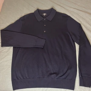 Mörkblå pikétröja från Uniqlo - Säljer en stilren mörkblå pikétröja från Uniqlo. Tröjan har långa ärmar och en klassisk krage med knappar framtill. Perfekt för en avslappnad men ändå uppklädd look.