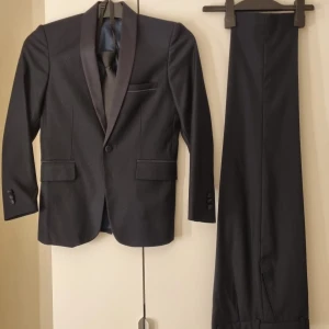 Svart kostym med väst och byxor - Elegant mörkblå kostym med kavaj, väst och byxor. Kavajen har en klassisk passform med två knappar och fickor. Västen matchar med knappar framtill och byxorna har en rak design. Perfekt för formella tillfällen.