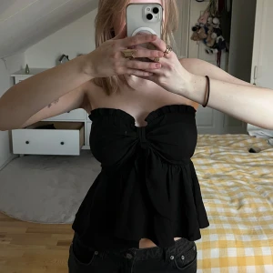 Svart axelbandslös topp - Snygg svart axelbandslös topp med volangdetaljer och knytning framtill. Perfekt för en stilren look.