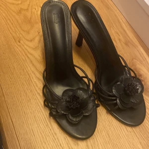 Svarta klackskor med blomdetalj - Eleganta svarta klackskor med en dekorativ blomma på ovansidan. Skorna har en smal klack och öppen tå, vilket ger en stilren look. Perfekta för att lyfta din outfit med en touch av elegans. Aldrig använda, endast testade och pris kan gärna diskuteras!