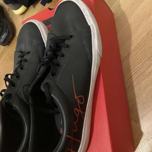 Svarta sneakers från Hugo - Snygga svarta sneakers från Hugo med vit sula och röd logga på sidan. Skorna har klassisk snörning och en stilren design som passar perfekt till vardags. De kommer i en röd originalkartong med Hugo-tryck. Helt nya Hugo boss skor  använda 1-2 gånger , boxen kommer med alltså som helt nya 