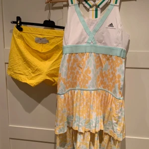 Mönstrad klänning från Adidas Stella McCartney - Säljer en fin klänning från Adidas Stella McCartney med ett blommigt mönster i gult och blått med tillhörande hot pants. Klänningen har en v-ringad design med mesh tyg och är ärmlös med en mintgrön detalj i midjan. Perfekt för en sportig look med stil. Stl 40 är mer (36/38 tycker jag lite mindre i storleken) Kan användas om du spelar tennis, Paddel  sommarfesten eller  beachen. Både klänning och de gula hotpants ingår i priset. 