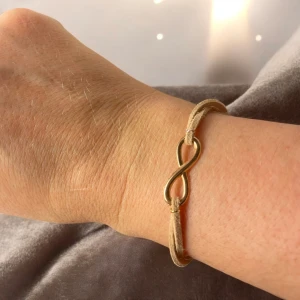 Beige armband med gulddetalj - Snyggt beige armband av konstlädersnöre, med en guldfärgad oändlighetssymbol i mitten. Armbandet har en enkel design med en justerbar rem och en liten karbinhake i guldfärg. Perfekt för att ge en subtil touch av elegans till din stil.