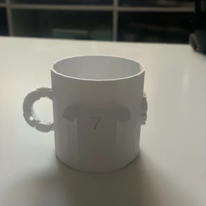 Vit mugg med fotbollstema - En vit mugg med ett fotbollstema. Muggen har ett handtag och är dekorerad med en tröja med nummer 7, samt texten 'Cristiano Ronaldo' inuti. Perfekt för fotbollsfans som vill ha en unik mugg.