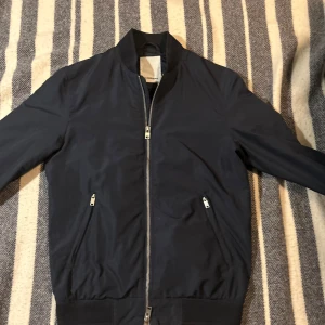 Mörkblå windbreaker från J.Lindeberg - Snygg mörkblå bomberjacka från J.Lindeberg med dragkedja framtill. Jackan har en klassisk bomberstil med ribbade muddar vid ärmslut och nederkant. Perfekt för en stilren look. Storleken är XS och inga defekter. Ifall du har några frågor är det bara att höra av sig👀👀