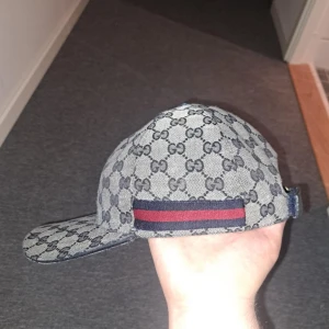 Gucci keps - Tar helst emot byten!!! Snygg keps från Gucci med det klassiska GG-mönstret i grått och svart. Kepsen är justerbar baktill och har en röd och blå randdetalj på sidan. Perfekt för att ge din outfit en lyxig touch.